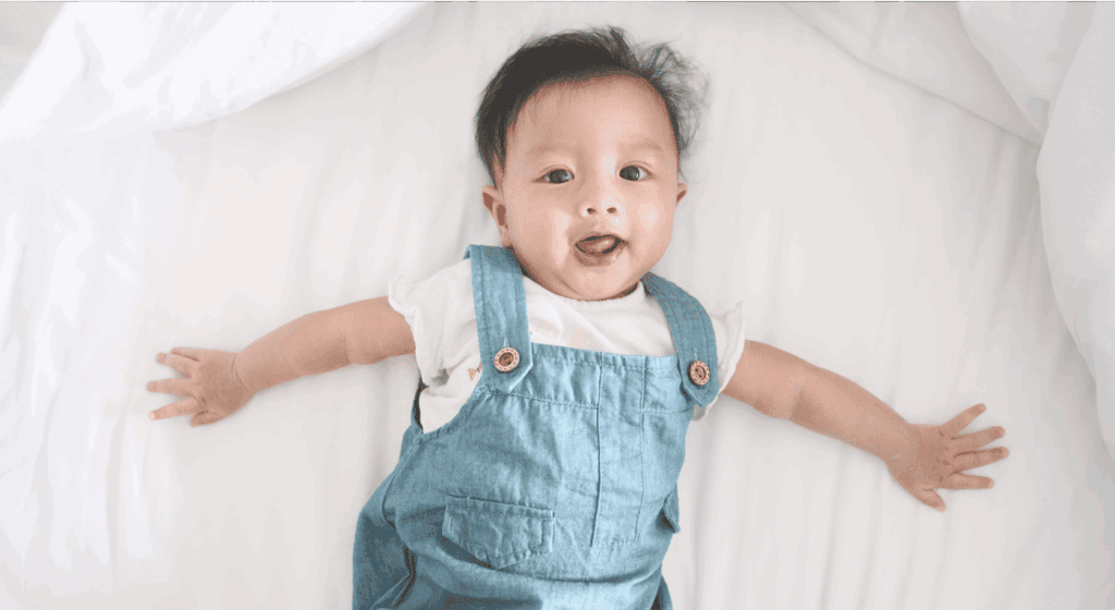 Happy baby lying on white bed, wearing denim dungarees, cheerful and playful.