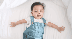 Happy baby lying on white bed, wearing denim dungarees, cheerful and playful.