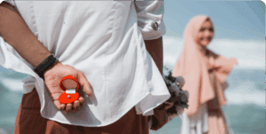Man holding engagement ring behind back on beach.