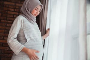 Pregnant woman in hijab standing by window.