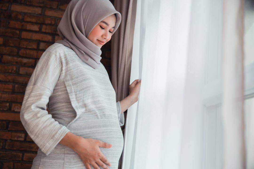 Pregnant woman in hijab standing by window.